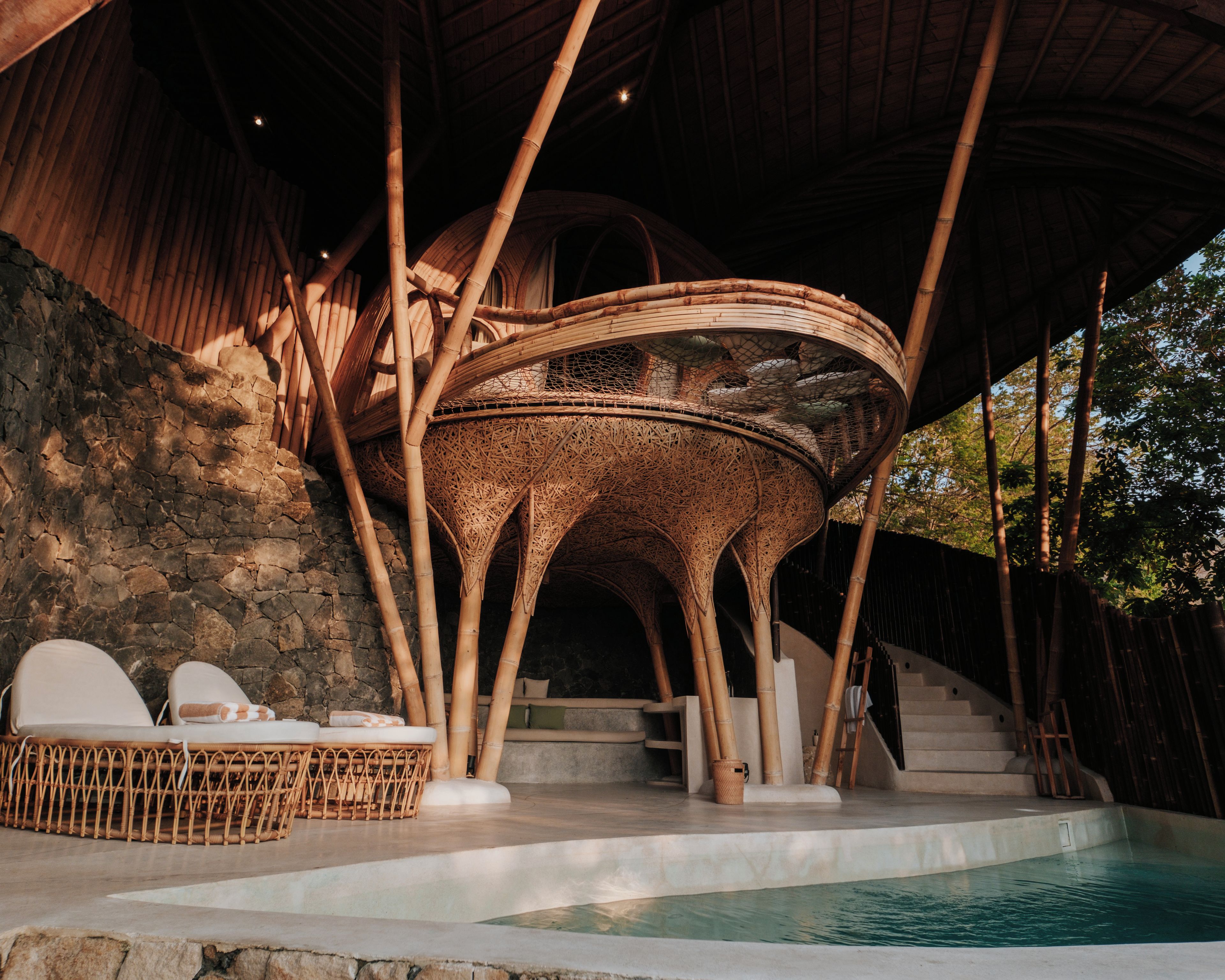 A pool area with a stone wall, lounge chairs, and a bamboo structure.