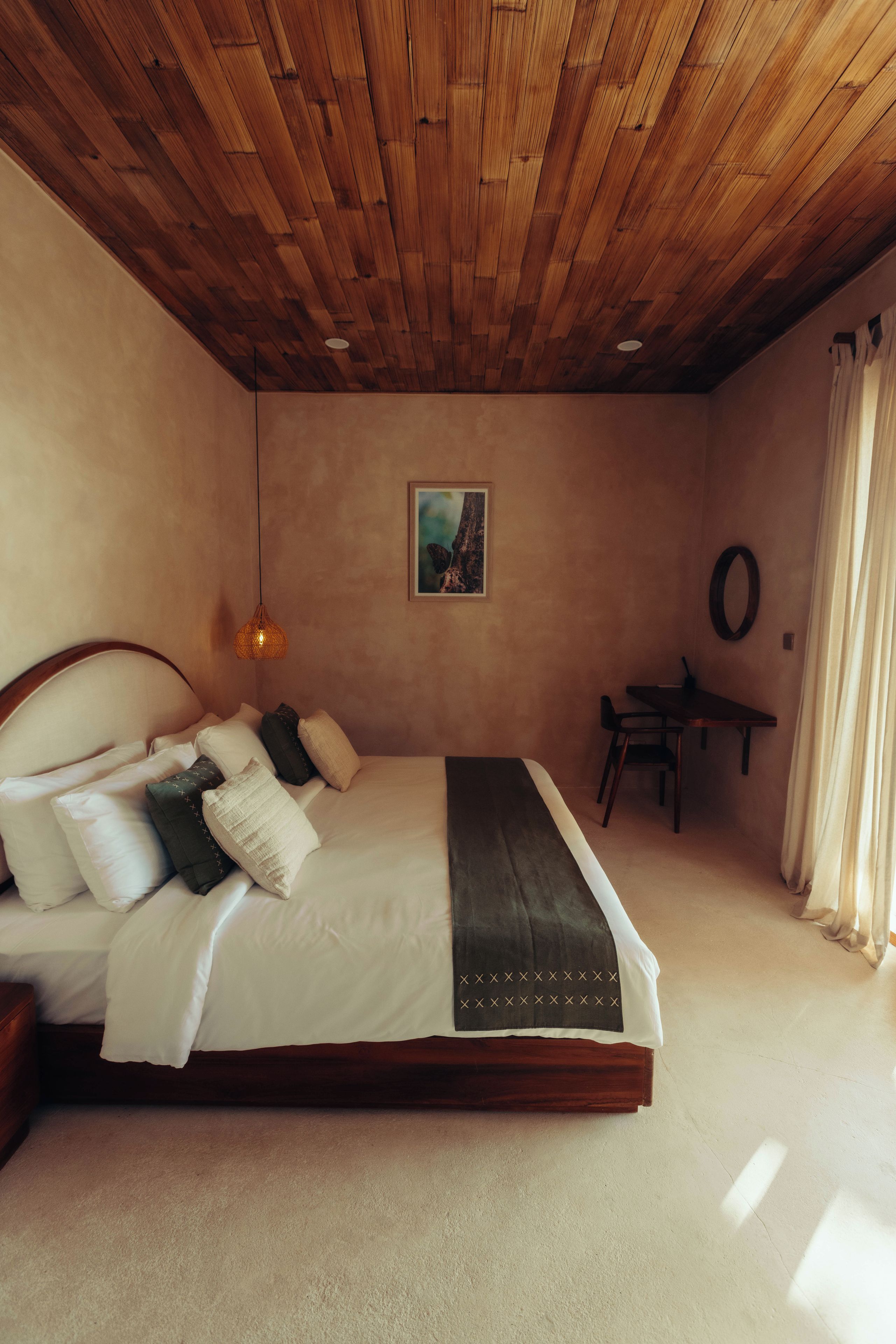 A bedroom with a wooden ceiling, a bed with multiple pillows, a bedside table, and a wall-mounted picture.