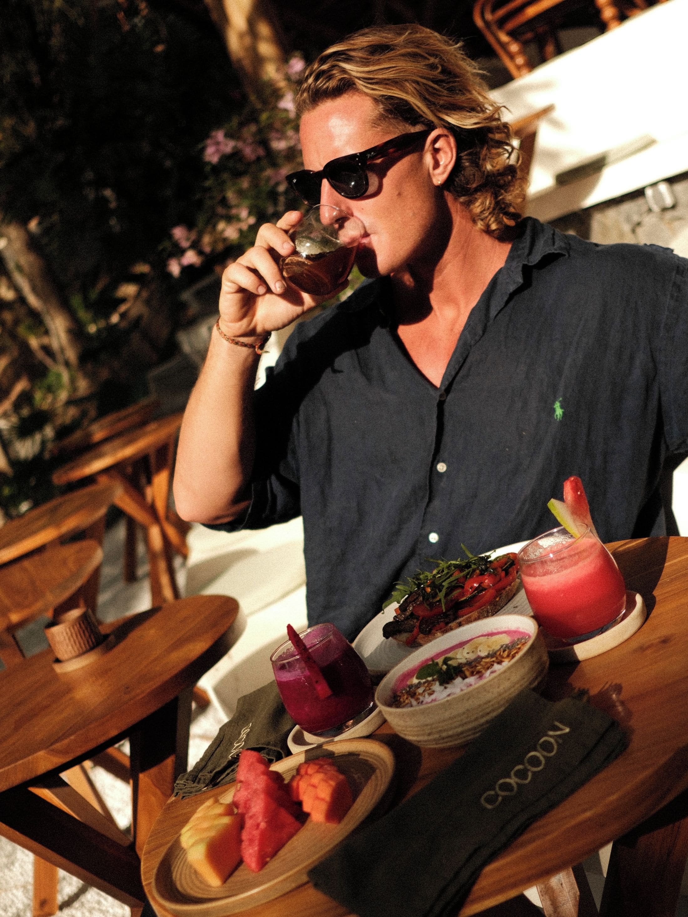 A person seated at a wooden table with a meal consisting of a salad, a bowl of soup, and a plate of watermelon.