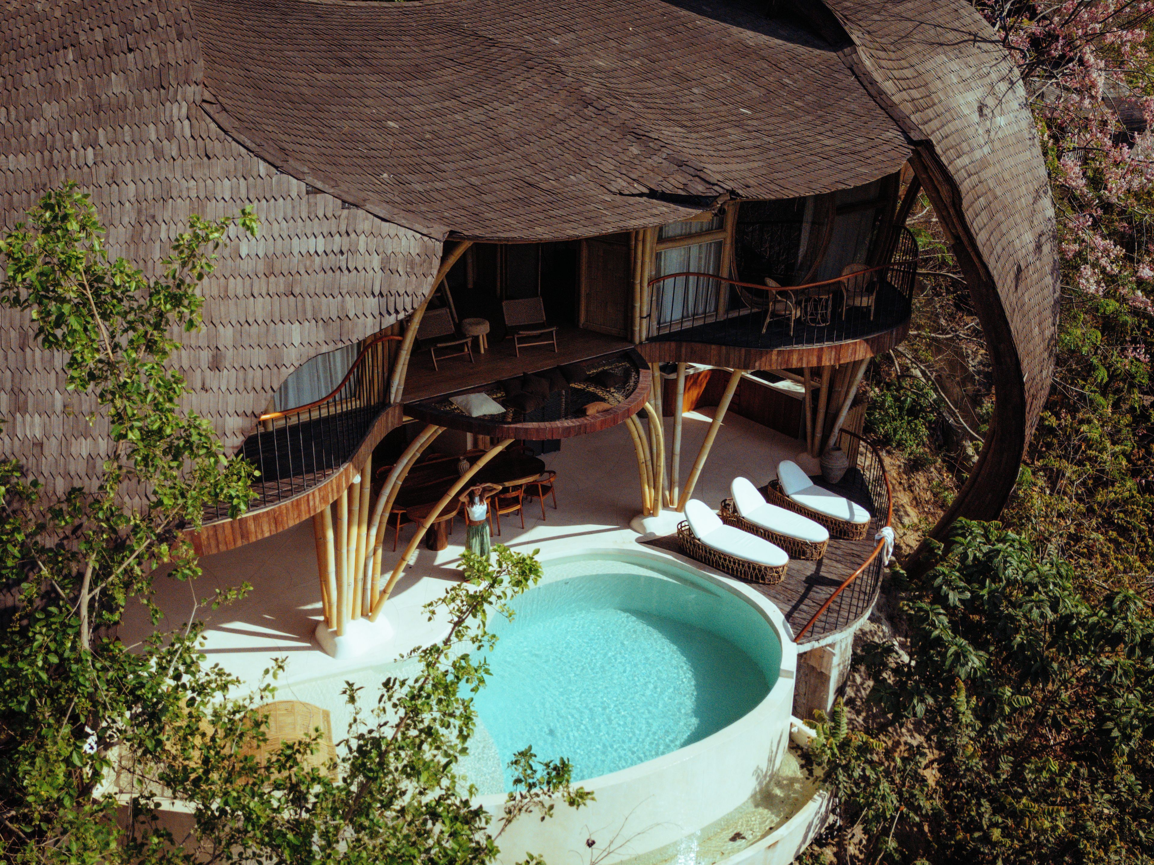 Un grand bain à remous circulaire, entouré d’une terrasse en bois, avec un espace salon couvert doté de chaises, et une vue sur la végétation environnante.