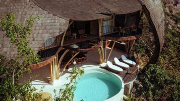 Un grand bain à remous circulaire, entouré d’une terrasse en bois, avec un espace salon couvert doté de chaises, et une vue sur la végétation environnante.