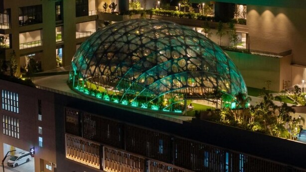 Ein modernes Gebäude mit einer großen, glockenförmigen Glasstruktur in der Mitte, umgeben von kleineren Gebäuden und Grünflächen.