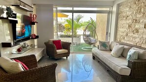Un salón moderno con mobiliario de mimbre, un televisor de pantalla plana y un amplio ventanal corredera que da a un patio con una silla colgante.