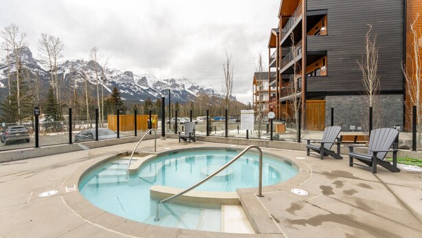 A pool area with a view of snow-capped mountains, surrounded by a building with balconies and a wooden deck. - Bell`s Butte | Pool, Hot Tub & Fitness | Parking (Canmore)