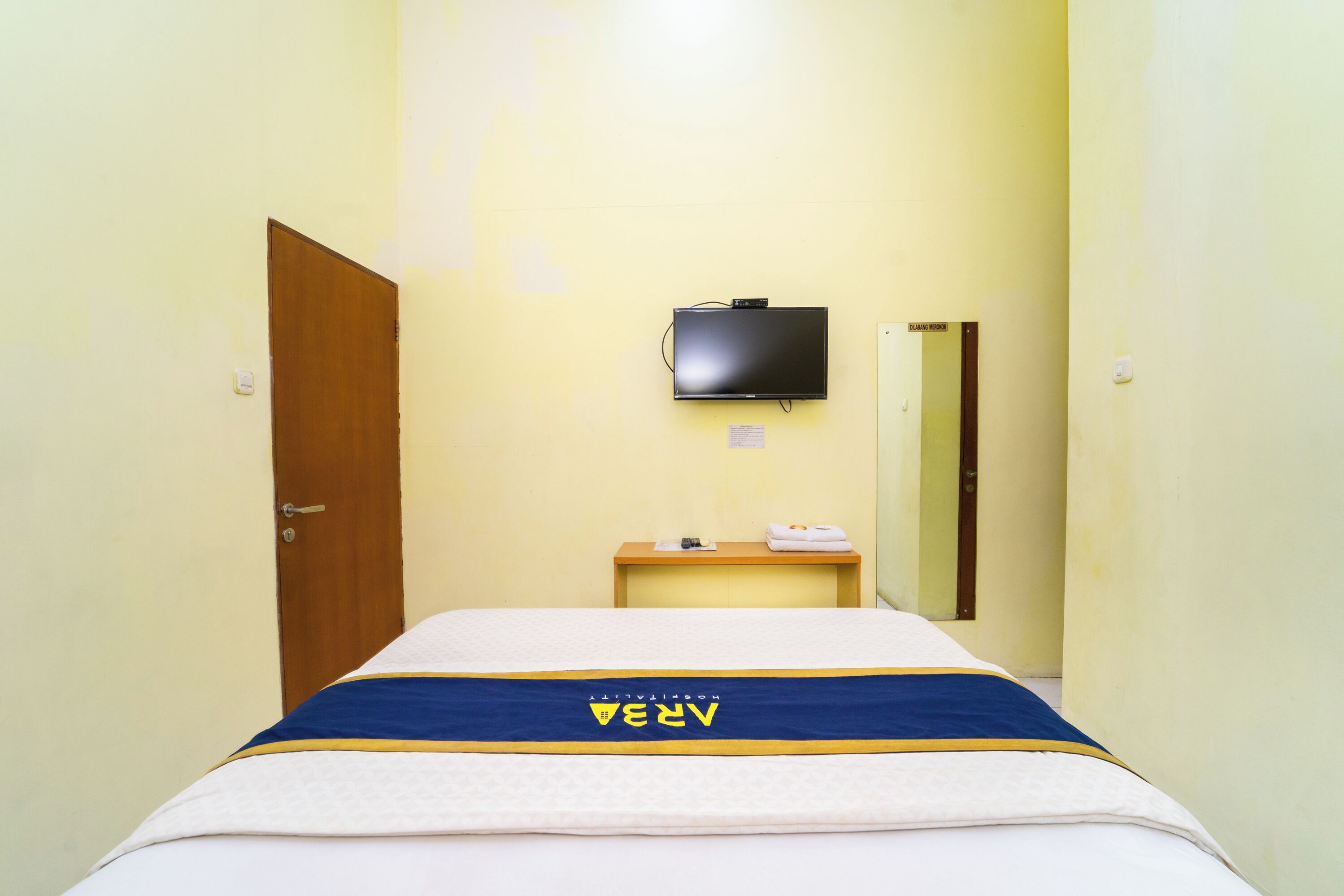 Una camera d'albergo con un letto, una TV a muro, un comodino in legno e uno specchio.