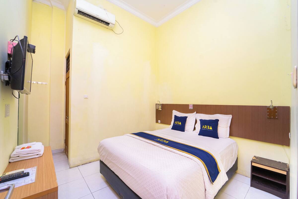 Ein Hotelzimmer mit Bett, Schreibtisch, Telefon, Unterlagen, Klimaanlage und einem an der Wand befestigten Fernseher.
