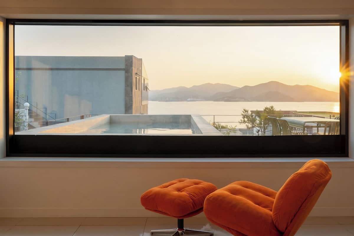 Una habitación moderna con un amplio ventanal que da a una piscina y montañas al atardecer.