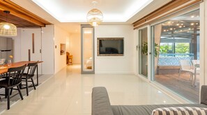 A modern living room with a flat-screen TV, a dining area with a wooden table and chairs, and a balcony with a pool. - Geoje Sea N Blue Family Pool Villa (Geoje)