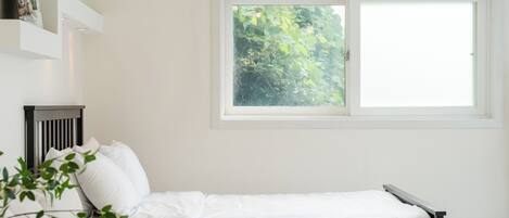 A bedroom with a bed, a window with a view of greenery, a radiator, and a wall-mounted shelf.