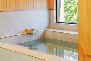 A modern bathroom with a glass bathtub, wooden paneling, and a window with a view of trees. - Gyeongju Sanmaru Pension (Gyeongju)