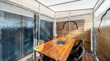 Un área de comedor al aire libre techada, con una mesa y sillas de madera, un lavamanos de metal y vistas a través de un muro de cristal.