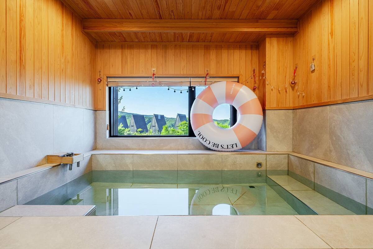 Un intérieur en bois avec un jacuzzi entouré de parois de verre, une fenêtre donnant sur des maisons et un anneau de sauvetage accroché au mur.