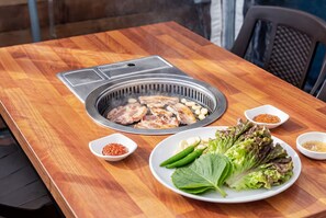 Een houten tafel met een ingebouwde grill, een bord vol groenten en kleine schaaltjes met sauzen.