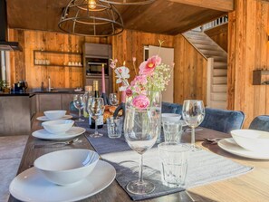 Un intérieur en bois avec une table à manger dressée pour quatre personnes, entourée de chaises, et un coin cuisine à l’arrière-plan.