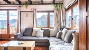Un salon chaleureux avec un canapé gris, une table basse en bois et de grandes fenêtres donnant sur un paysage montagneux.