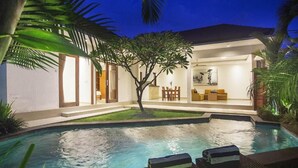 Una piscina con sillas de descanso, un área de comedor y un árbol en el centro.