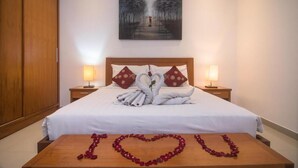 Una habitación de hotel con una cama bien hecha, dos lámparas de noche, un cuadro en la pared y un arreglo de pétalos de rosa con forma de corazón sobre la mesa.