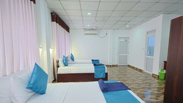 Una habitación de hotel con tres camas, cada una con ropa de cama azul y blanca, un ventilador de techo, un equipo de aire acondicionado y piso de baldosas.