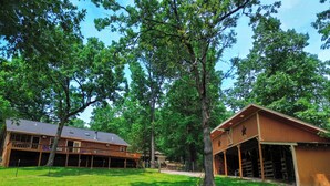 A two-story wooden cabin with a balcony, surrounded by lush green trees and a well-maintained lawn. - Rustic Retreat near Norfork Lake (Clarkridge)
