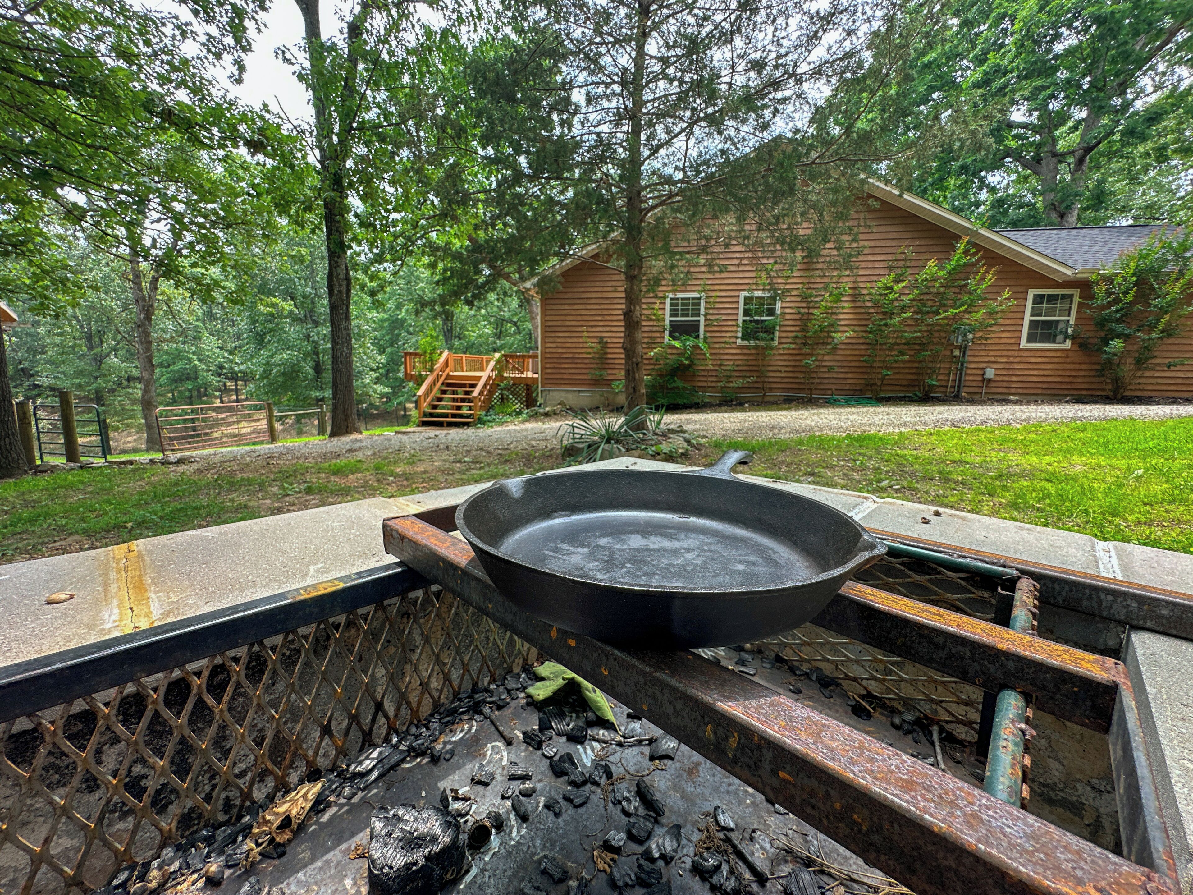 A rustic outdoor grill with a cast iron pan, surrounded by a wooden deck and a wooden house in a wooded area.