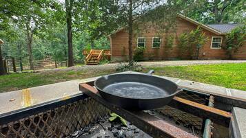 A rustic outdoor grill with a cast iron pan, surrounded by a wooden deck and a wooden house in a wooded area.