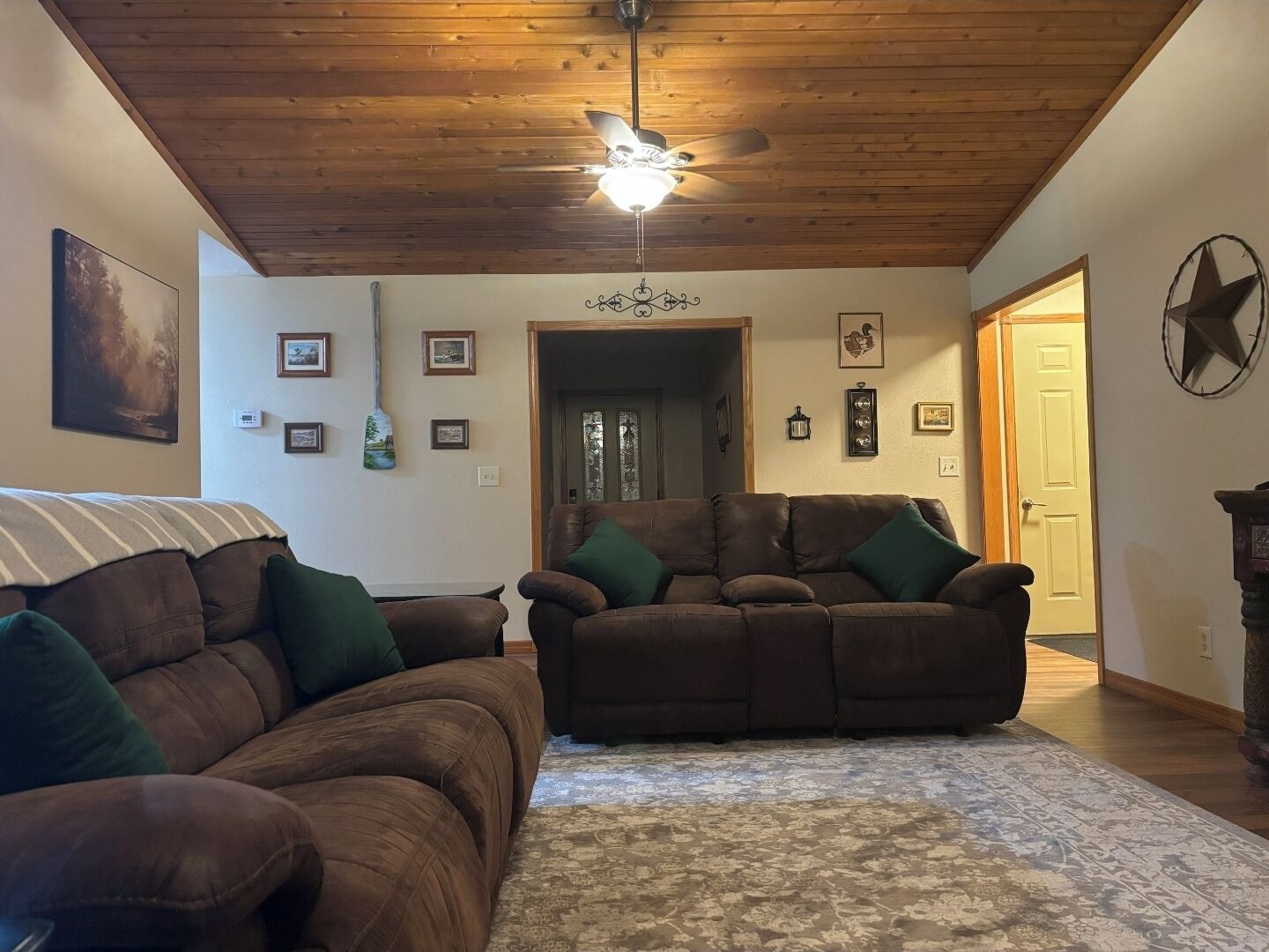 A living room with two brown leather sofas, a patterned rug, wooden ceiling, and a door leading to another room.