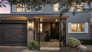 Une maison d’habitation avec une porte de garage sombre, une entrée bien éclairée et un petit jardin agrémenté de rochers et de plantes.