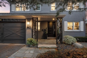 A residential house with a dark garage door, a well-lit entrance, and a small garden area with rocks and plants. - Experience luxury living at lovely renovated home (Toronto)