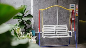 A white swing chair with a yellow, red, and blue frame in a room with a grey wall and a potted plant. - Kangsar Inn 73 (George Town)
