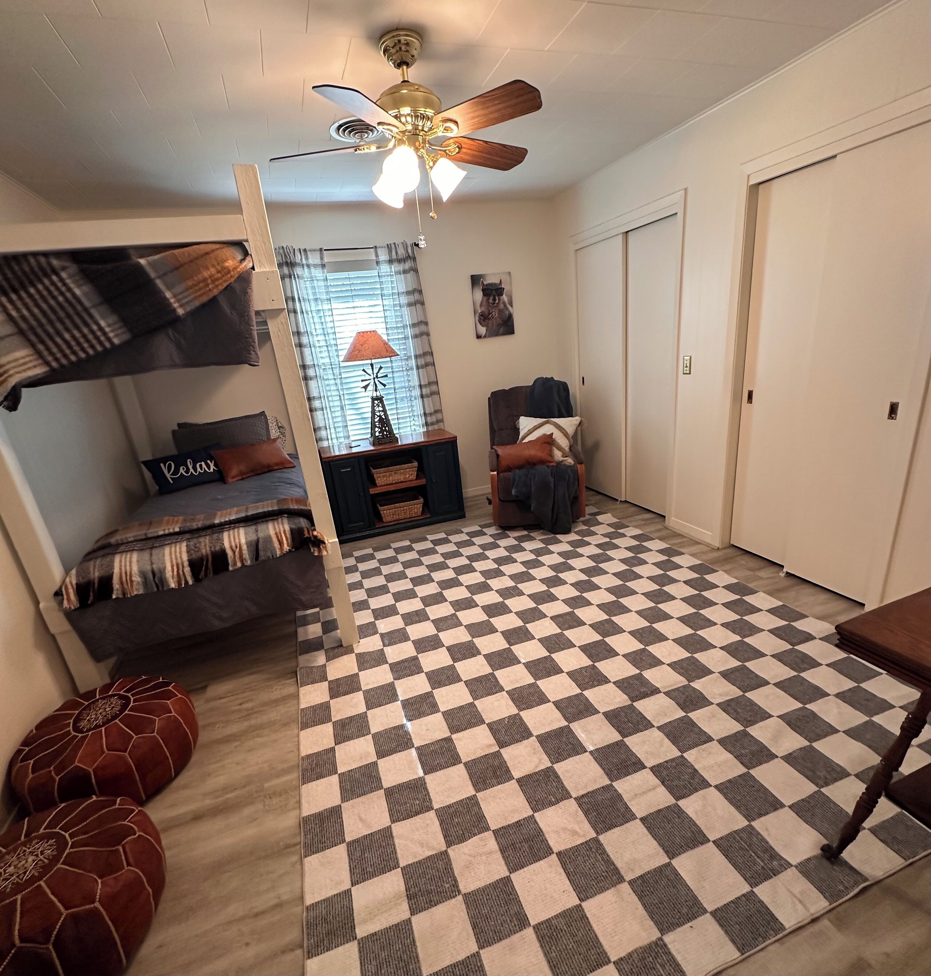 A room with a checkered floor, a ceiling fan, a bed with a blanket, a chair, and a door.