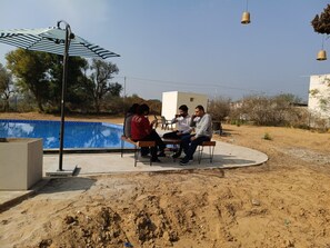 A group of people seated around a table outdoors, near a swimming pool, with a large umbrella and hanging lights in the background. - Vardhan Resorts (Nawalgarh)