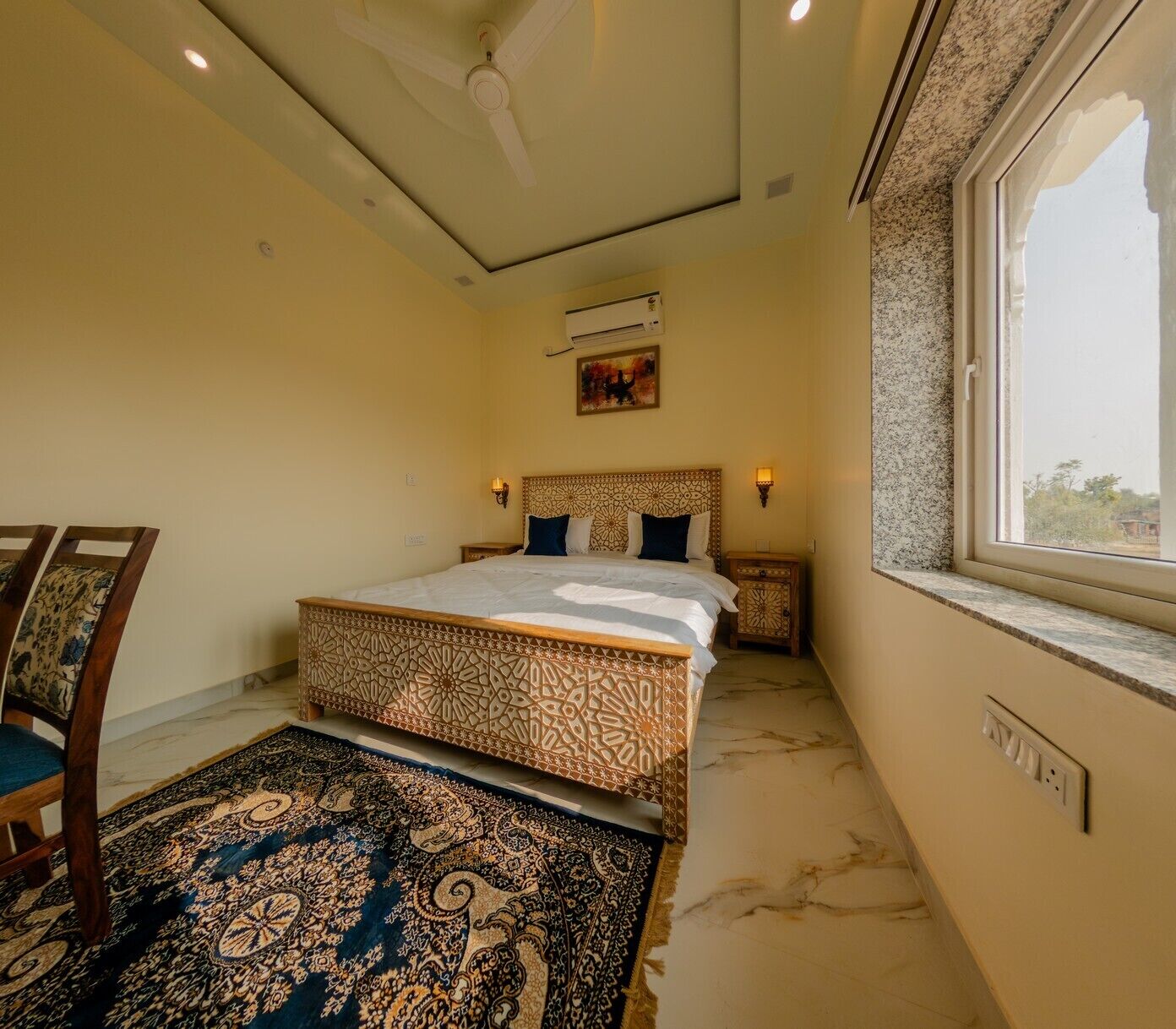A bedroom with a bed, a nightstand, a chair, a patterned rug, a ceiling fan, an air conditioning unit, a window with a view, and a wall-mounted light fixture.