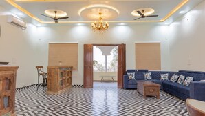 A spacious living room with a patterned floor, a blue sofa set, a chandelier, ceiling fans, and a view through a doorway. - Vardhan Resorts (Nawalgarh)