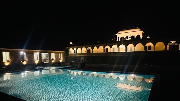 A swimming pool with a tiled floor and a building with arched windows in the background. - Vardhan Resorts (Nawalgarh)