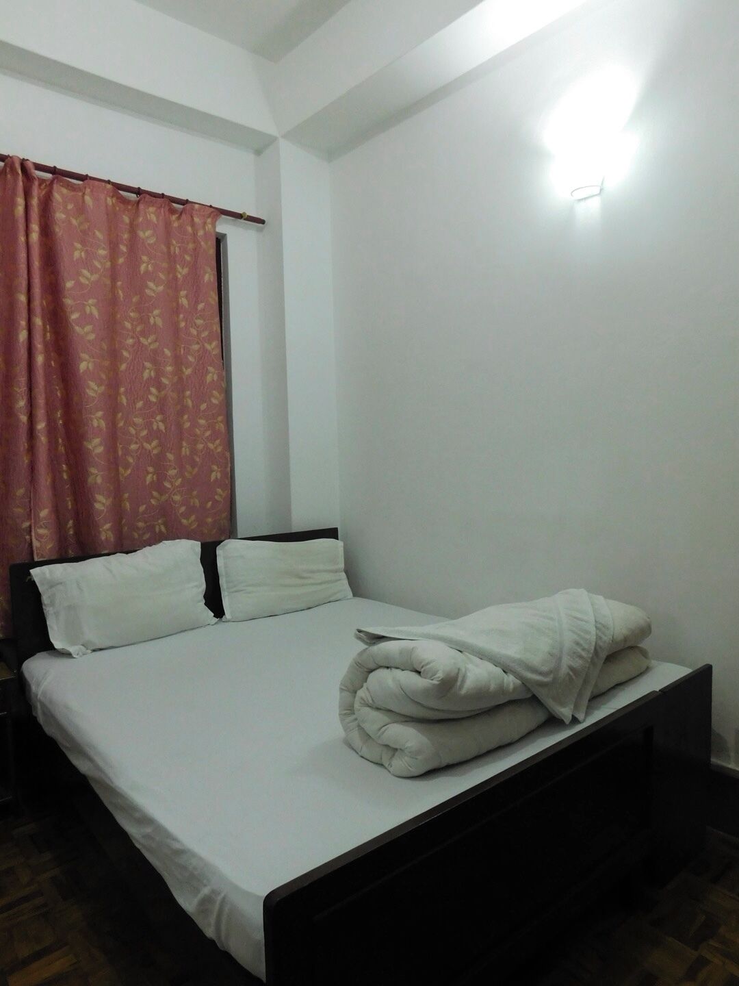 Una cama individual con ropa blanca de cama y una manta doblada, un cabecero de madera, una mesita de noche, una cortina roja con estampado y un aplique de pared.