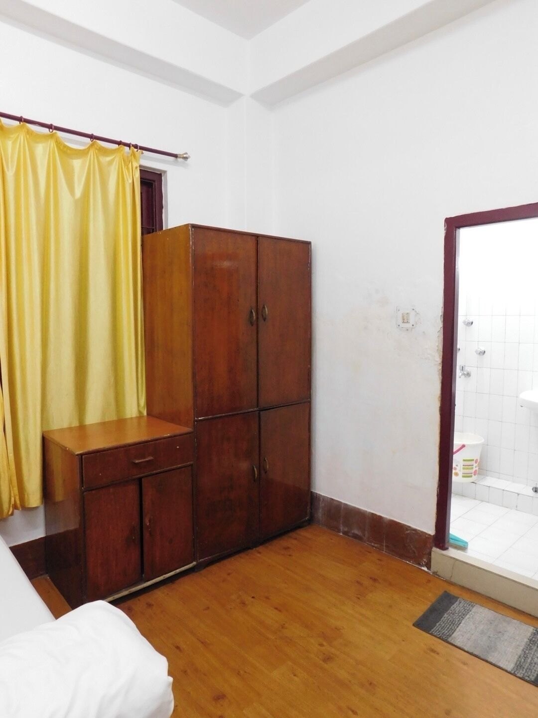 A room with a wooden wardrobe, a bed, and a bathroom with a white sink and tiled walls.