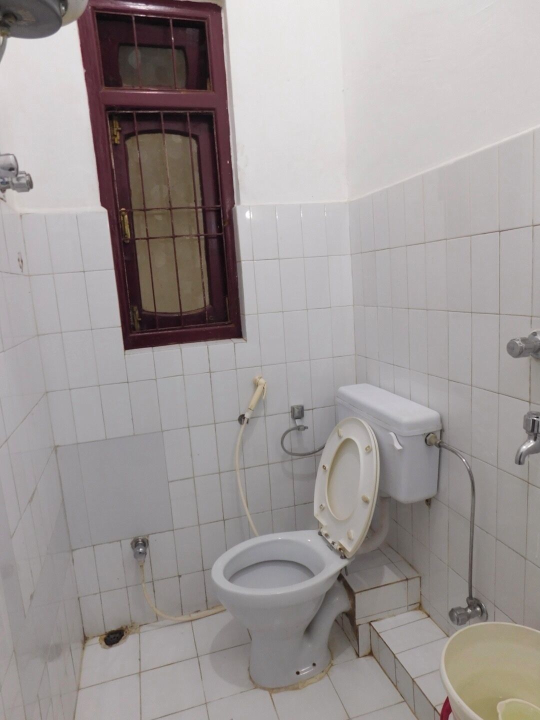 A small bathroom with a toilet, a window with a metal grille, and a bucket on the floor.