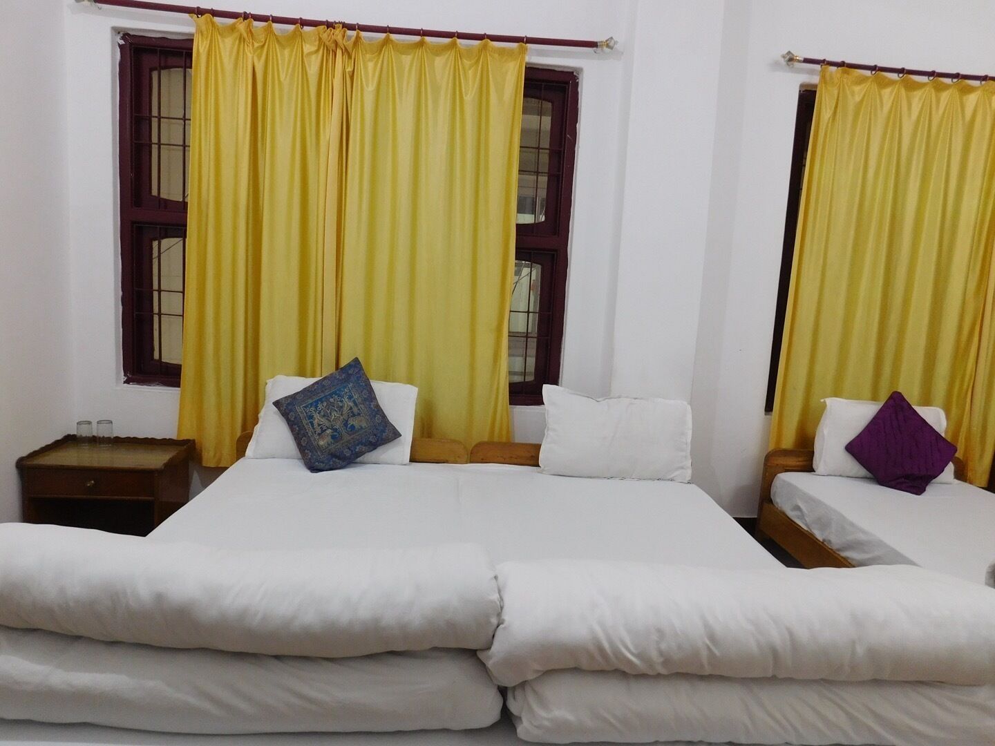 A room with two beds, each with white bedding and a single pillow. There is a nightstand with a lamp on the left side of the beds.