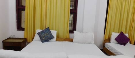 A room with two beds, each with white bedding and a single pillow. There is a nightstand with a lamp on the left side of the beds.