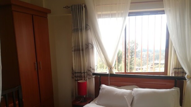 A bedroom with a bed, a wardrobe, a bedside table with a lamp, and a window with curtains. - Becca Hotel Meru (Meru)
