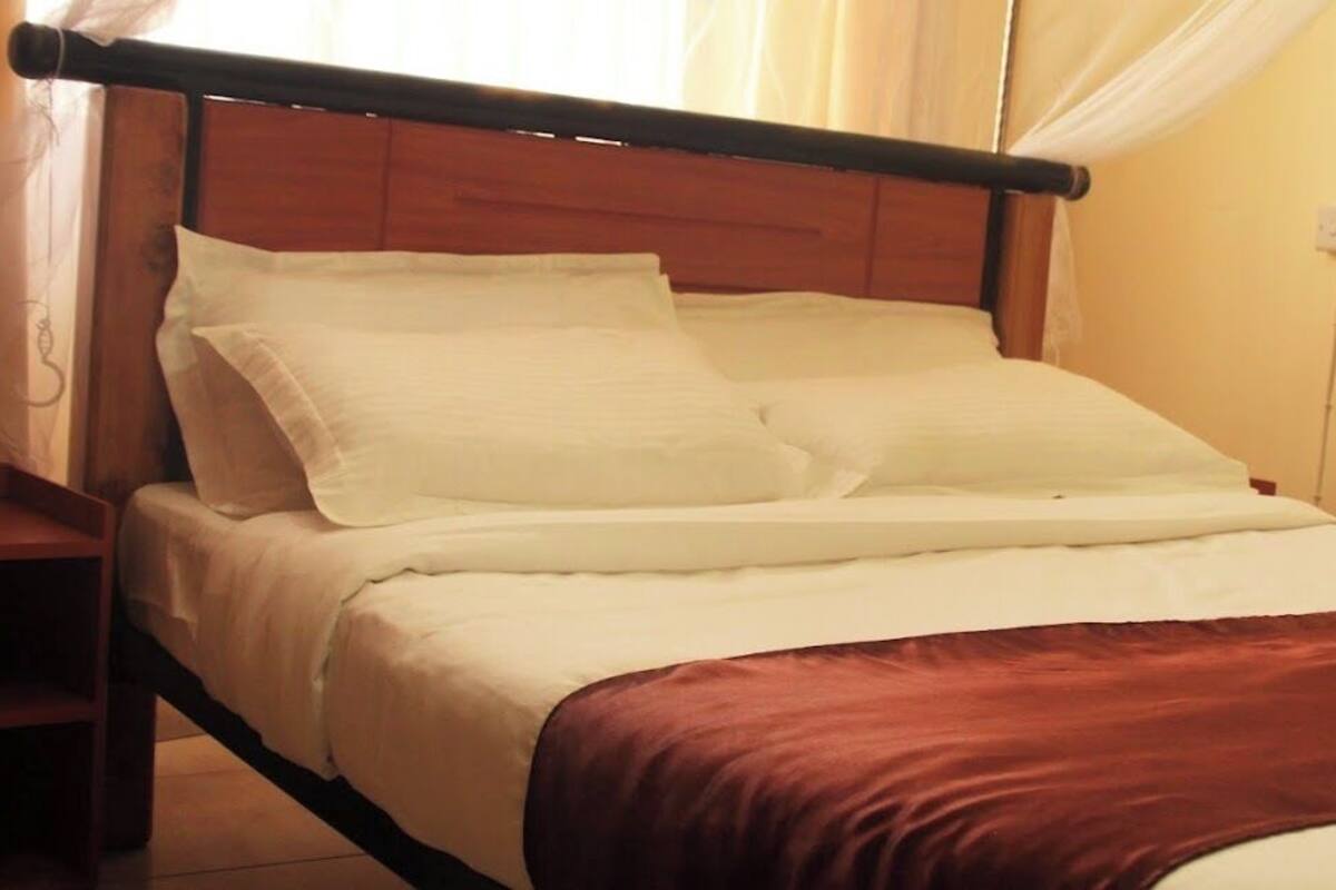 A neatly made bed with white linens and a brown throw blanket, a wooden headboard, and a nightstand with a lamp.