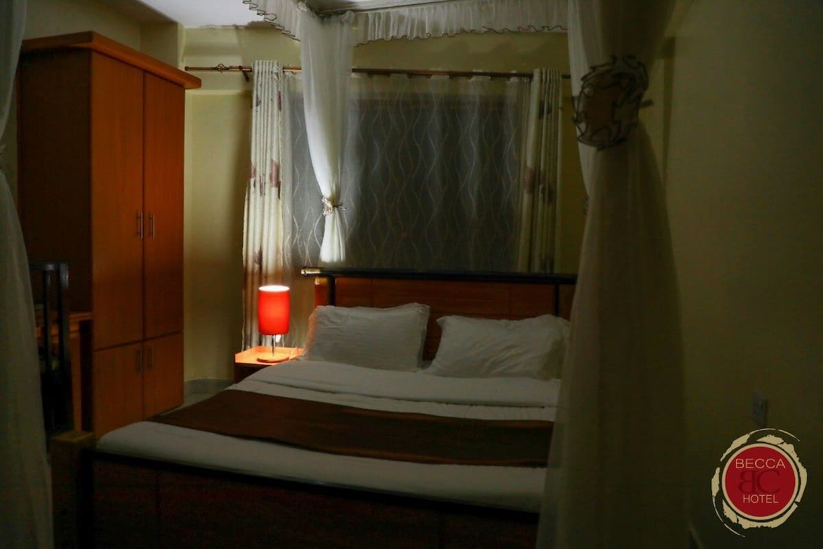 A hotel room with a bed, a nightstand with a red lamp, a wooden wardrobe, and a window with curtains.