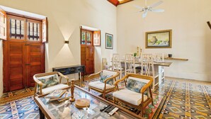 Uma sala de estar ampla, com mesa de centro de vidro, cadeiras de madeira, ventoinha no teto e piso estampado.