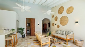 Uma sala de estar ampla com piso xadrez, um sofá, uma mesa de centro e um conjunto de jantar. Há ventiladores de teto, luminárias fixadas na parede e um espelho grande.