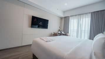 A hotel room with a large bed, a flat-screen TV mounted on the wall, a small table with a chair, and a window with curtains.