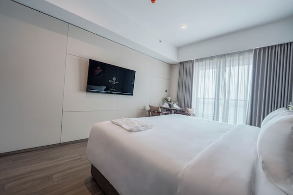 A hotel room with a large bed, a flat-screen TV mounted on the wall, a small table with a chair, and a window with curtains.