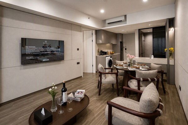 A modern living room with a flat-screen TV, a dining area with a table set for two, and a kitchen in the background.