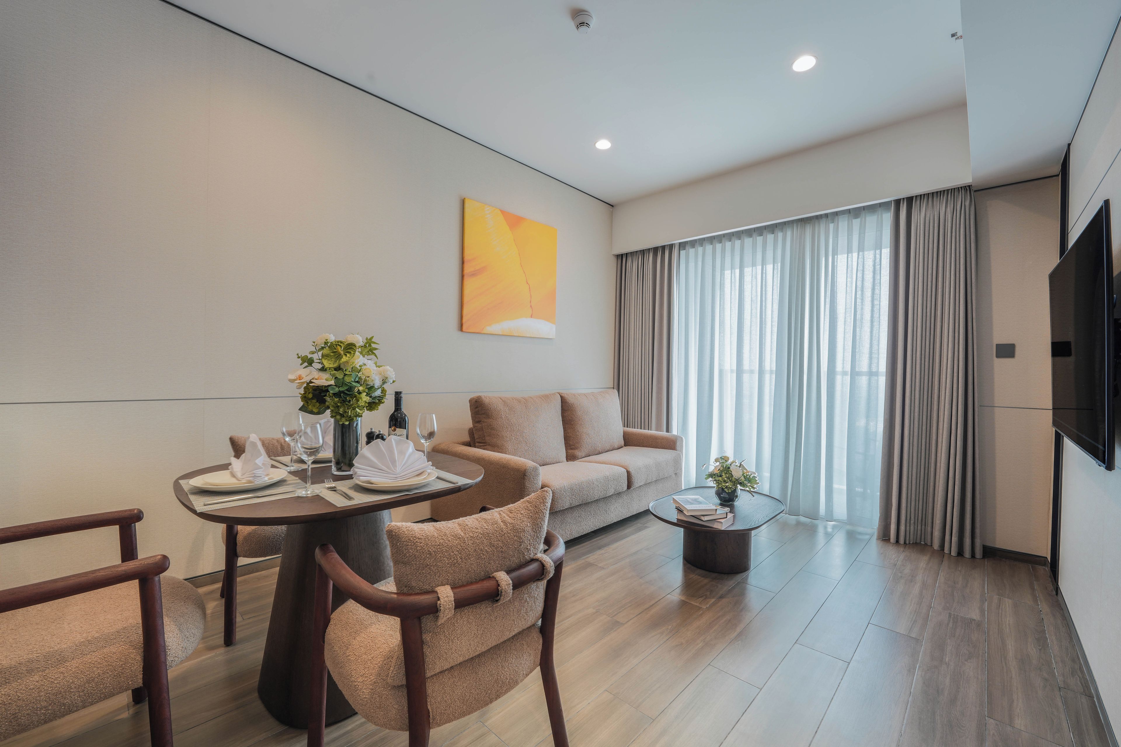A modern hotel room with a dining area, a sofa, a TV, and a large window with curtains.