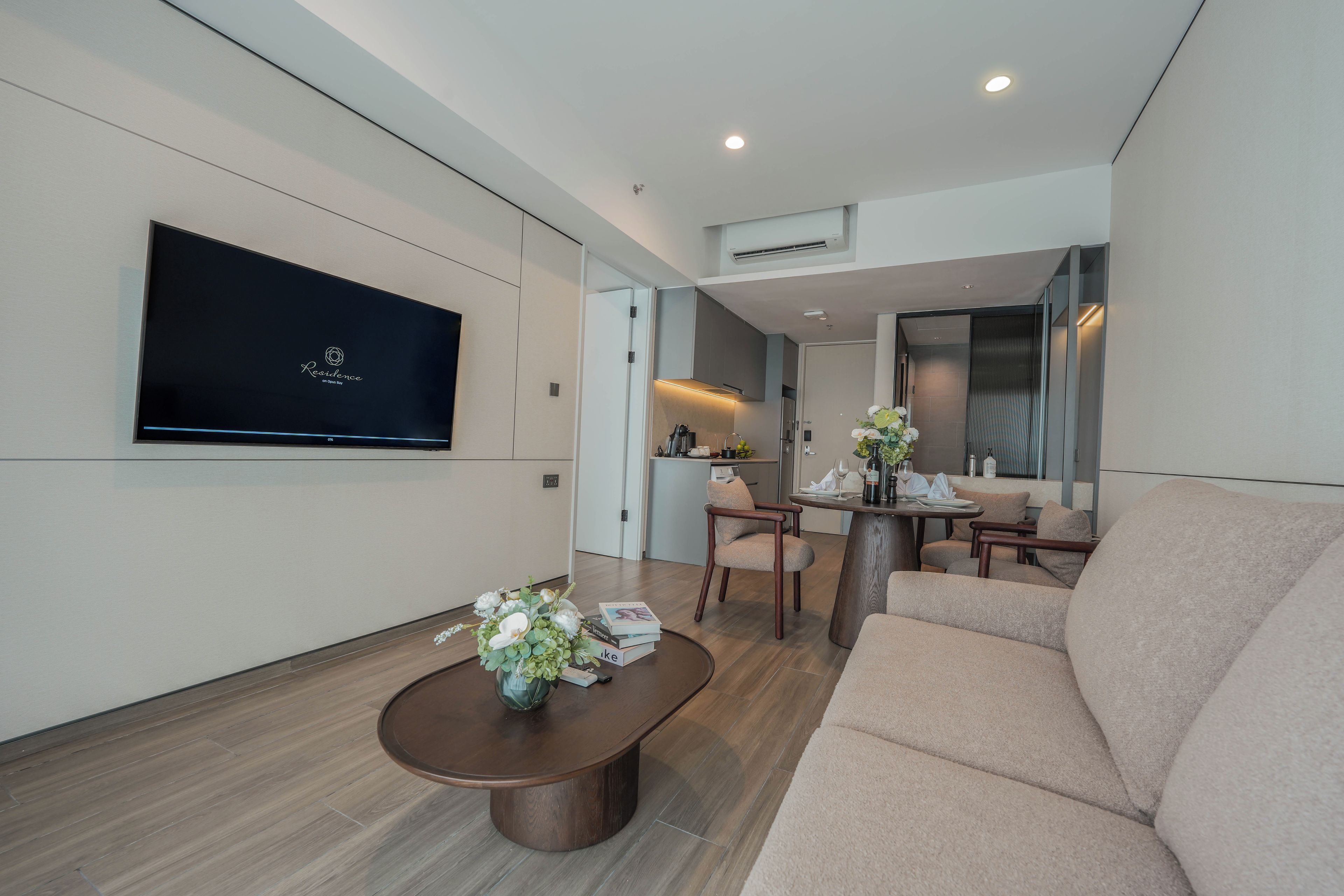 A modern living room with a flat-screen TV mounted on the wall, a round wooden coffee table with a floral centerpiece, and a beige sofa.