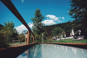 Una piscina con cubierta de madera, rodeada de exuberante follaje y un cielo azul despejado.